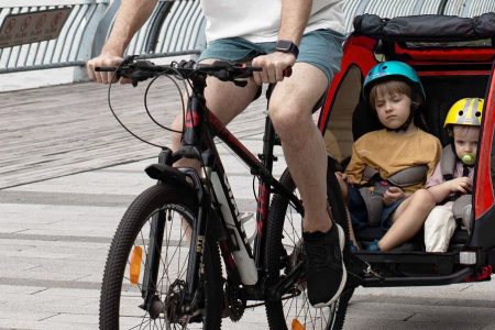 duas crianças dentro de um reboque de bicicleta infantil sendo puxadas pela bicicleta do pai