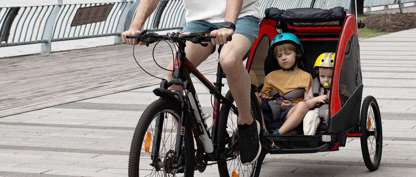 duas crianças dentro de um reboque de bicicleta infantil sendo puxadas pela bicicleta do pai