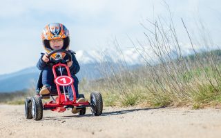 menino com um capacete laranja andando com um kart a pedais vermelho no campo