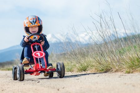 menino com um capacete laranja andando com um kart a pedais vermelho no campo