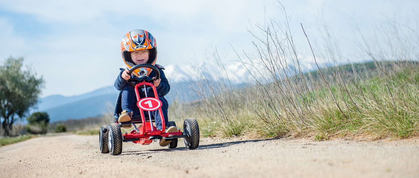 menino com um capacete laranja andando com um kart a pedais vermelho no campo