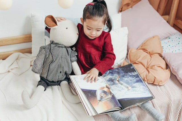 menina lendo um livro infantil ao lado do seu urso de pelúcia