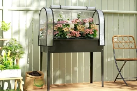 Floreira elevada com cobertura transparente, rodeada de plantas e flores num terraço de madeira.